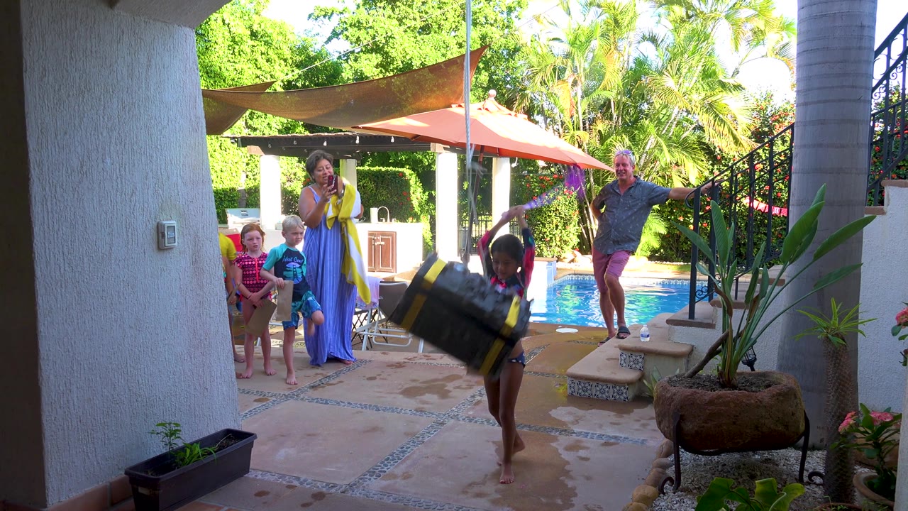 A child with a purple stick reaches toward the large tire swing as children and adults watch from beside the pool, with bright greenery and shade fabric behind them.