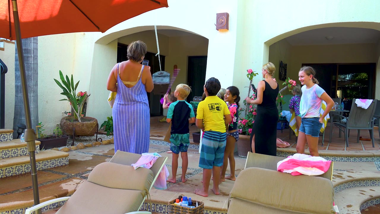 The San Jose Del Cabo patio scene shows adults and children near a dangling water toy, with a red umbrella overhead and outdoor furniture under arched openings behind them.