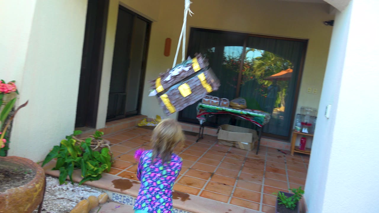 In San Jose Del Cabo, a child in a purple patterned shirt faces a hanging brown piñata while swinging a pink stick on a terracotta patio with plants and sliding doors behind.