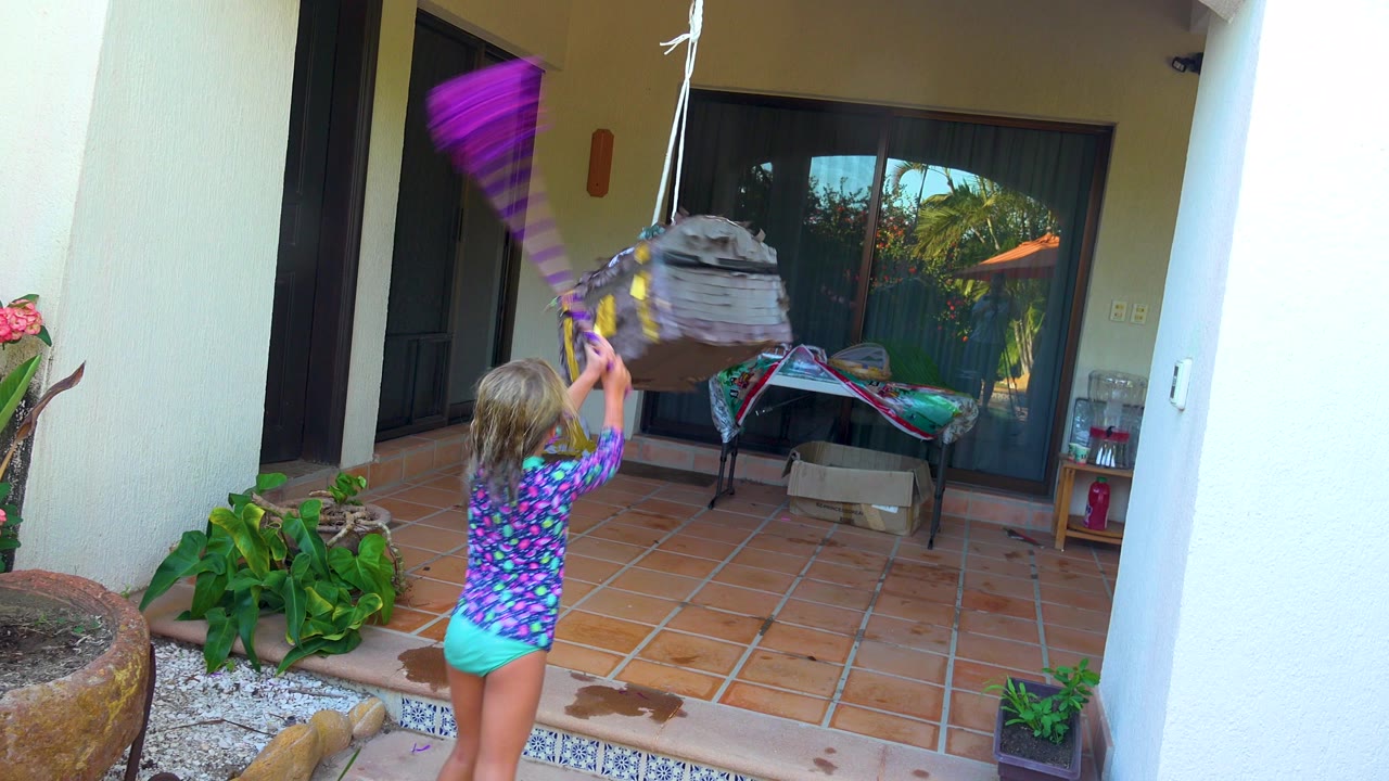 In San Jose Del Cabo, Mexico, a child in a colorful shirt swings a purple stick upward at a gray hanging piñata beside potted plants on a covered tiled patio.