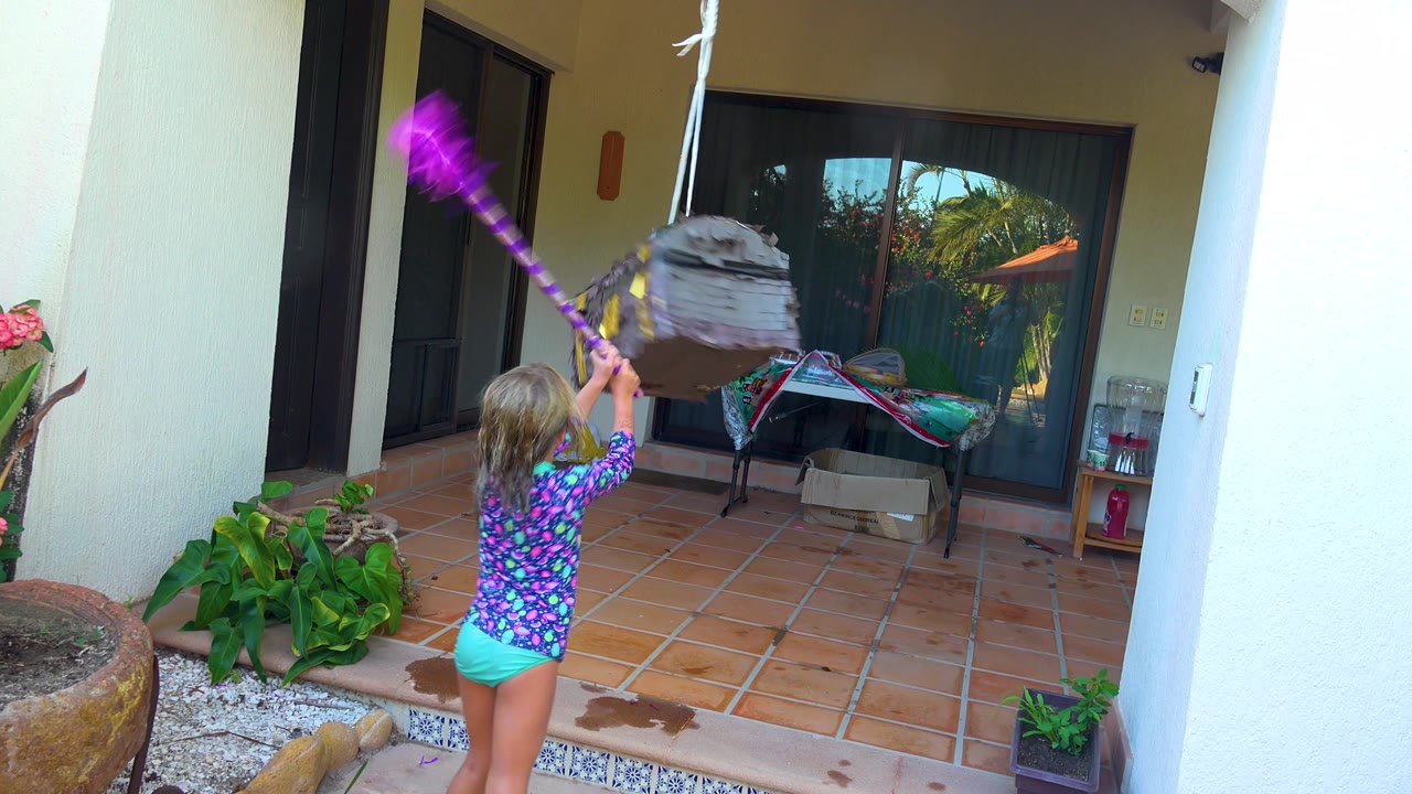 Child reaches up with a purple stick as the piñata hangs in front of sliding glass doors.
