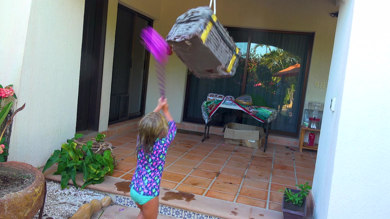 In San Jose Del Cabo, Mexico, the gray piñata swings high toward the upper right while the child raises the purple stick from below on the tiled patio.