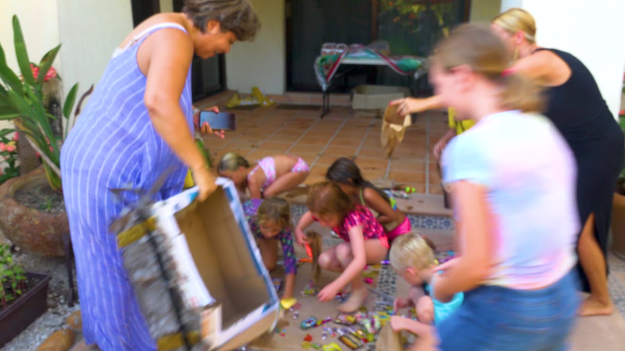 In San Jose Del Cabo, adults lean over a tiled patio as children crouch and reach for scattered colorful treats near potted plants and a house entry.
