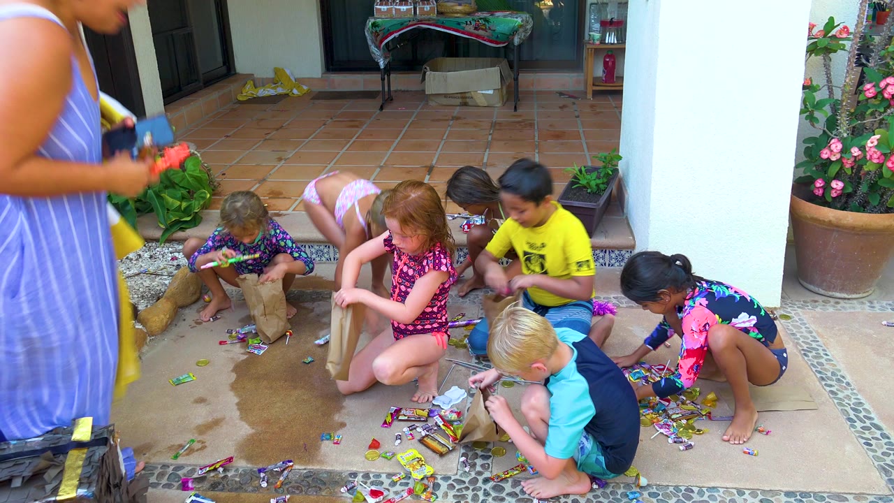At 0:00 in San Jose Del Cabo, several children in swimwear crouch on a wet tiled patio picking up candy while an adult in a blue striped dress is partly visible at left.