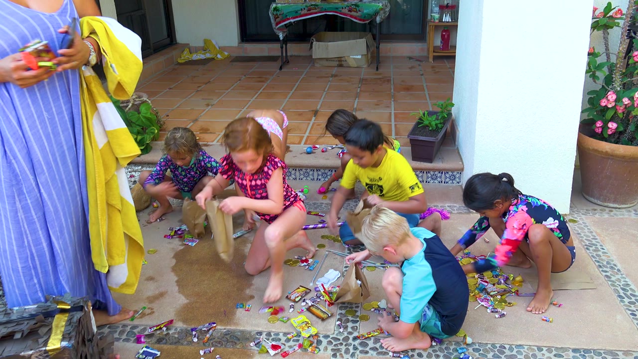 At 0:02 in San Jose Del Cabo, an adult holding a yellow towel and small object stands in the foreground while children crouch behind her to collect candy from the patio.