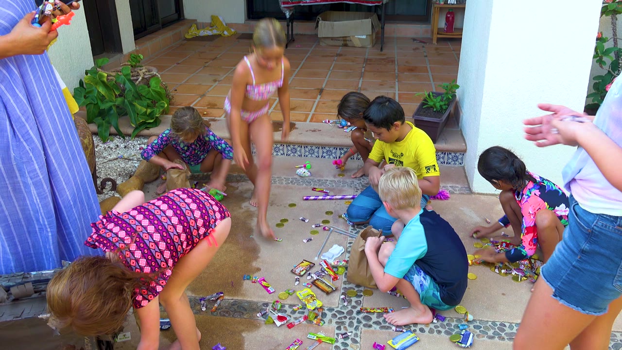 Children in swimwear and T-shirts collect scattered candies on a tiled patio in San Jose Del Cabo, Mexico, while adults are partly visible at both edges of the frame.