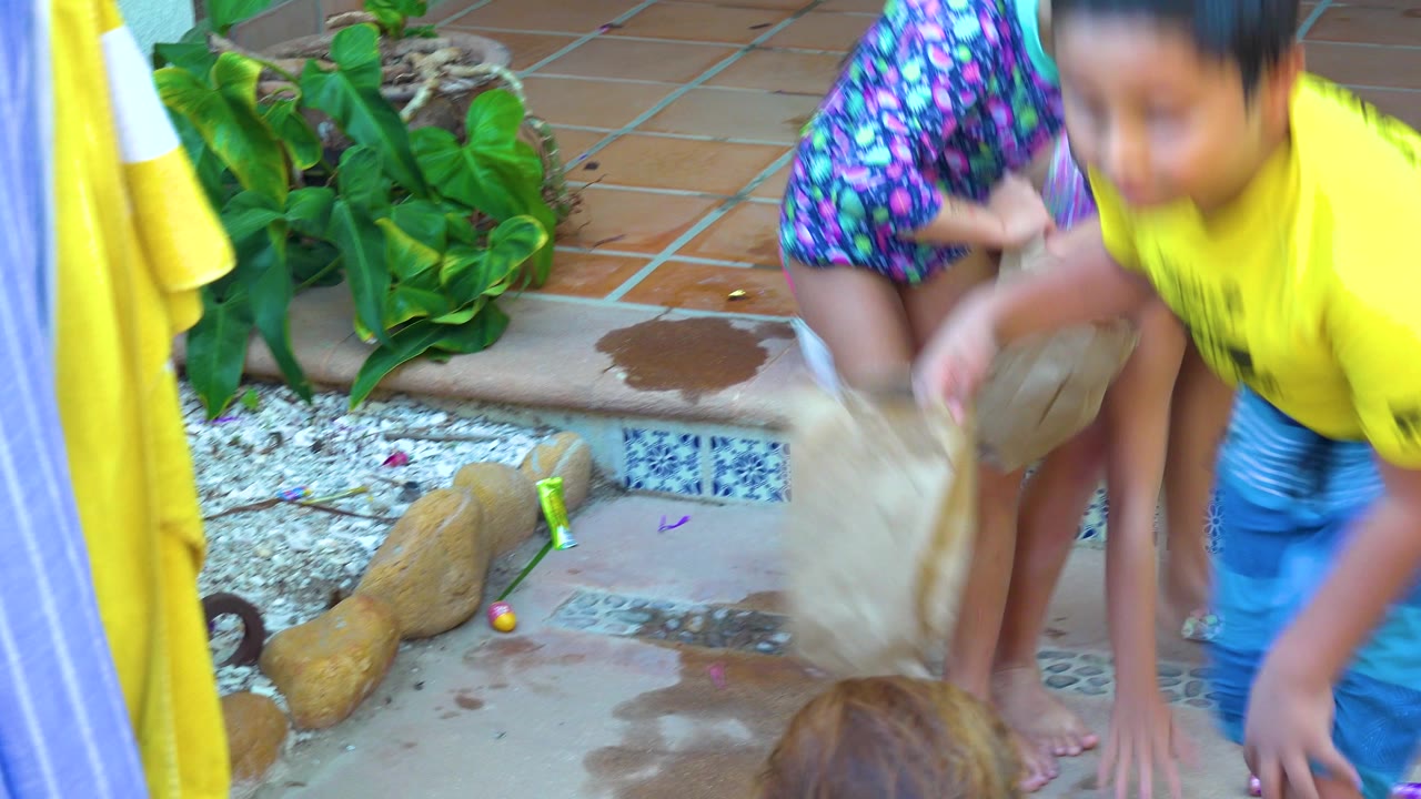 In San Jose Del Cabo, children lean over paper bags on a wet tiled patio while plants and patio steps sit behind them.