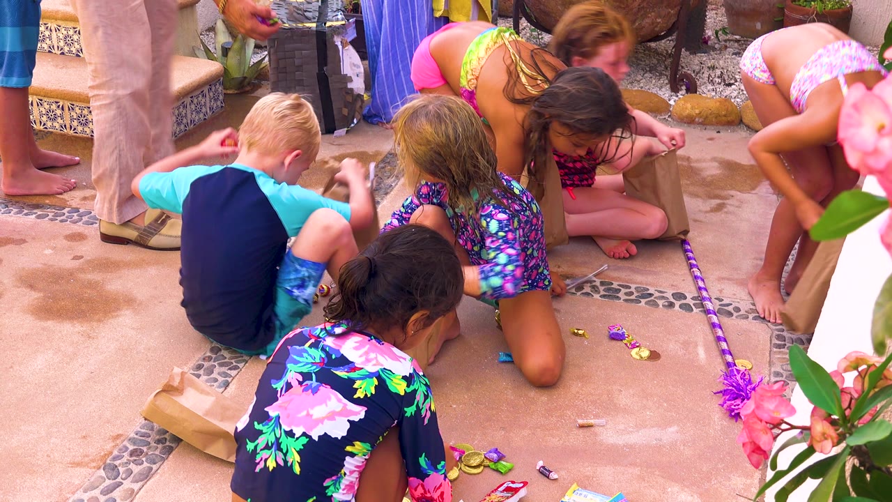 Children in bright swimsuits reach toward small items on the tiled patio in San Jose del Cabo, Mexico, while adults stand at the edges of the group.