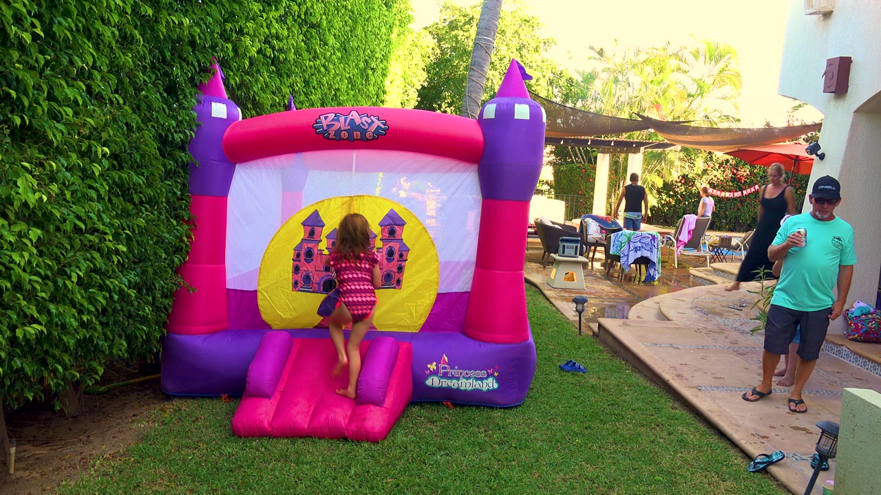 A child steps up onto the pink inflatable ramp, centered on the castle graphic, while adults and patio seating remain visible in the backyard setting.