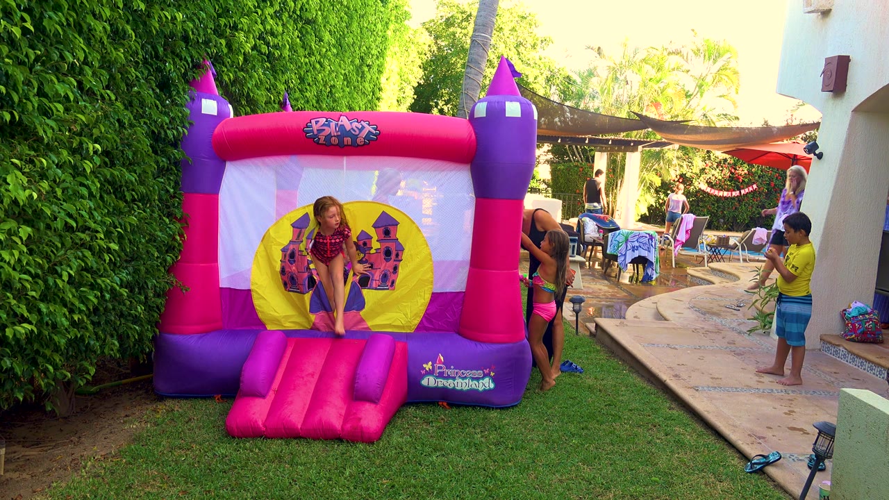 A girl stands on the front step of a pink and purple inflatable bouncy castle in a San Jose del Cabo backyard, with adults and children near a patio to the right.
