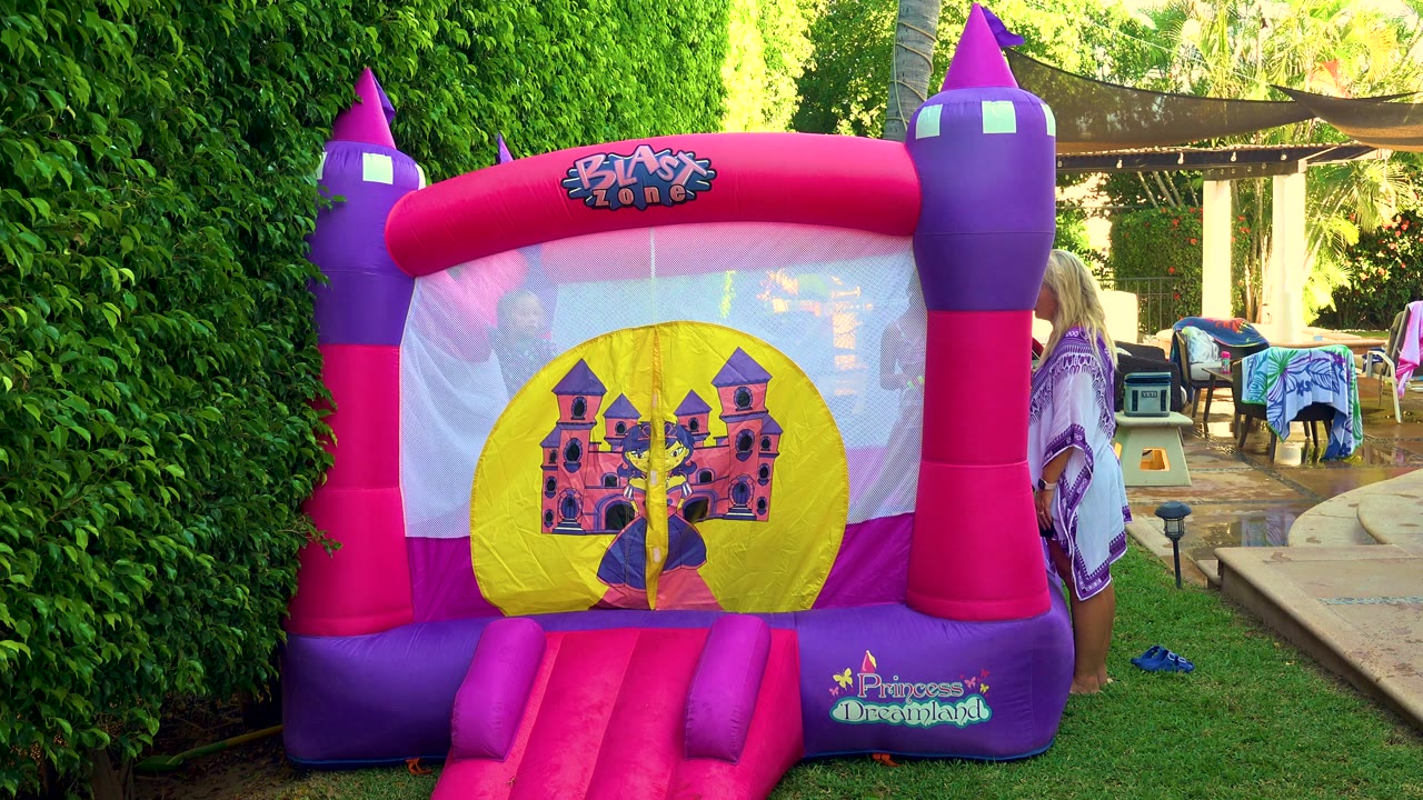Pink and purple princess-themed bouncy castle in a San Jose del Cabo backyard, with green hedges, grass, patio furniture, and a partial adult figure at the right edge.
