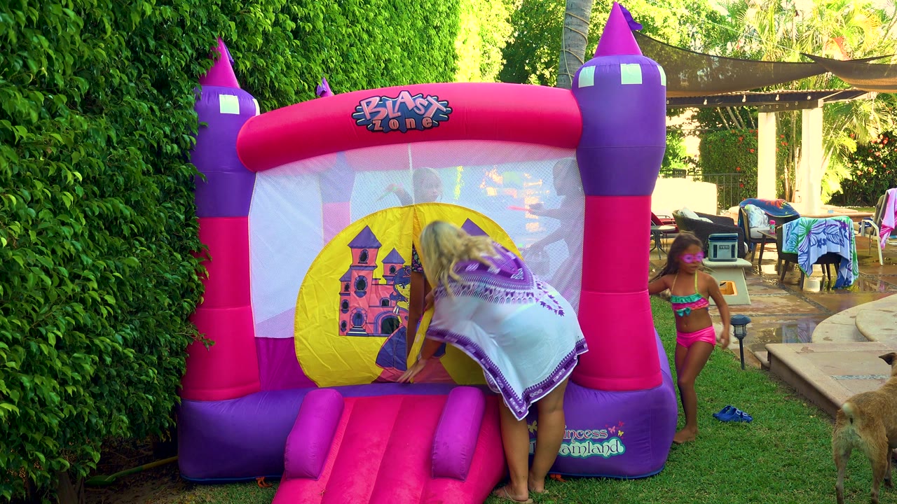 In San Jose Del Cabo, a girl covered by a towel bends into the yellow entrance of a pink and purple bouncy castle while another child in pink stands beside the yard.