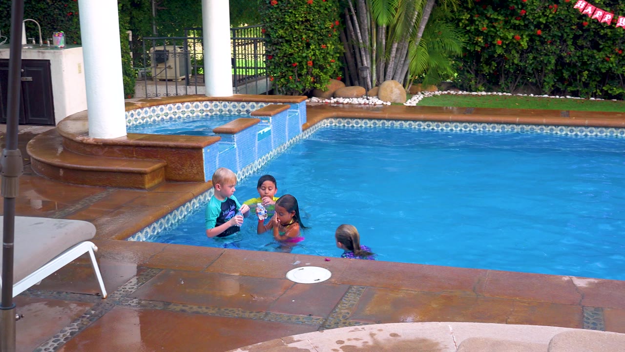 Children gather near the edge of a residential swimming pool in San Jose Del Cabo, with blue water, wet tile, pool steps, white columns, and greenery behind them.