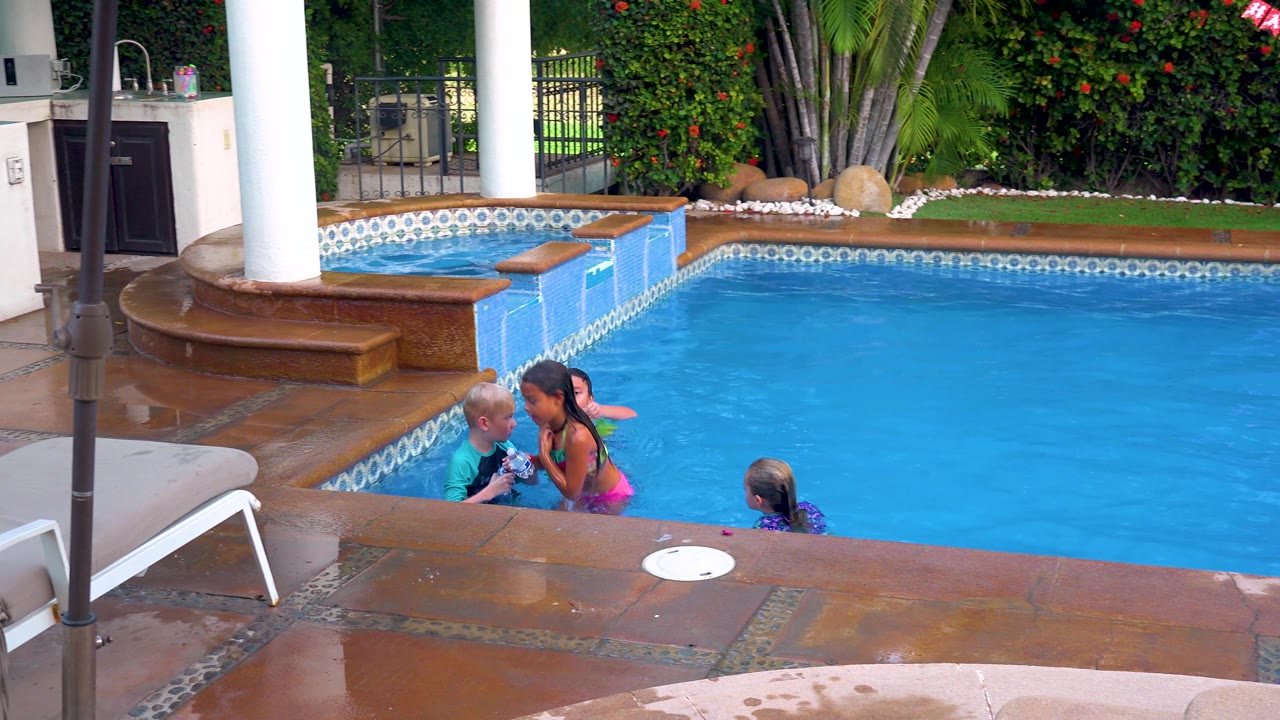 Two children lean close near the pool wall in the San Jose Del Cabo backyard pool while another child stays in the water to the right, with the patio and hedge framing the scene.