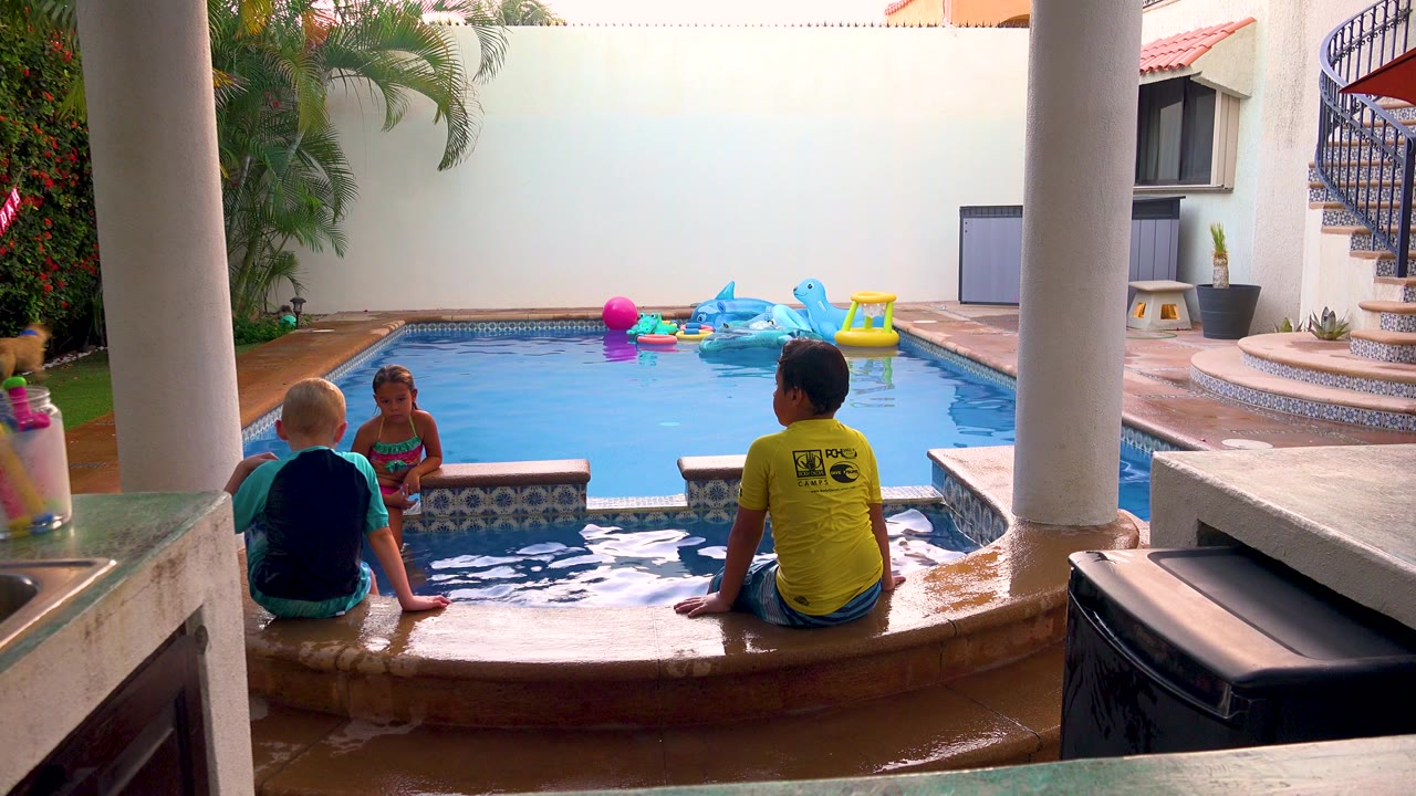 At 0:00 in San Jose del Cabo, Mexico, three children sit on the pool coping beneath patio columns while colorful inflatable toys float in the blue water behind them.