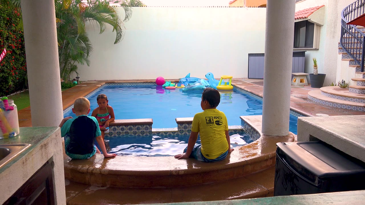 At 0:02, the children stay near the pool edge as ripples catch the light and a spiral staircase, tiled steps, and pool inflatables define the enclosed San Jose del Cabo backyard.