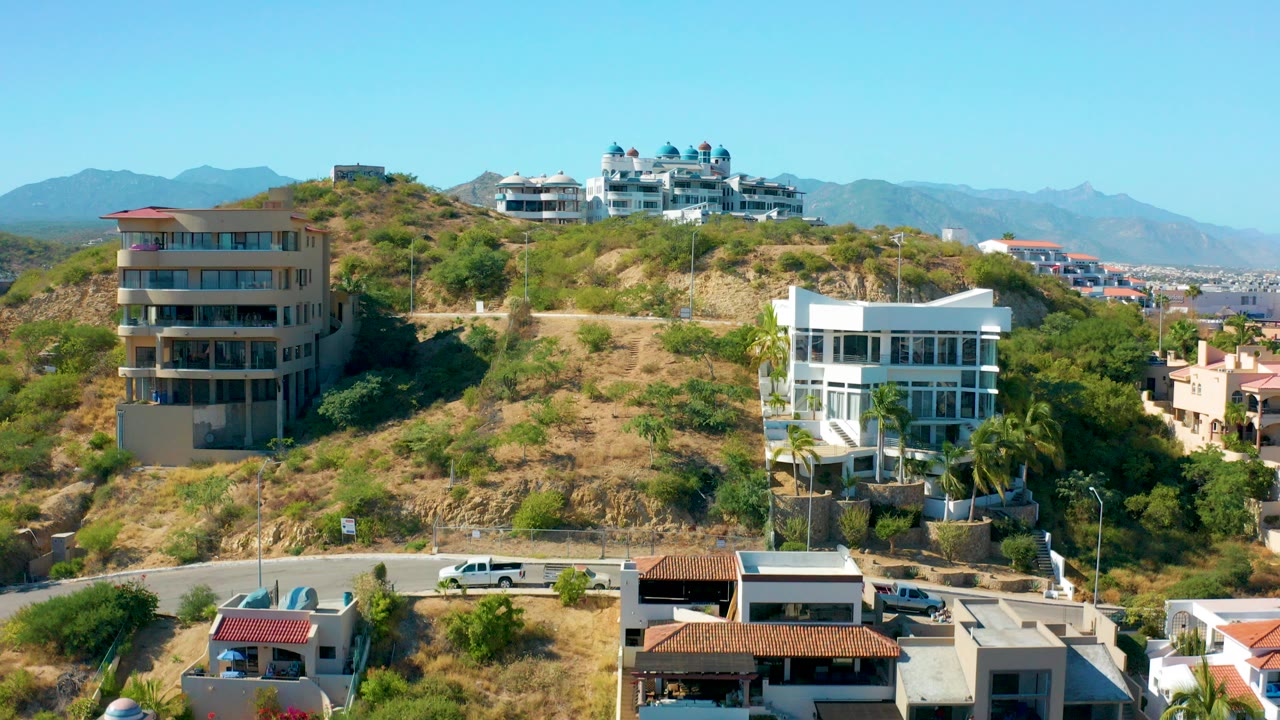 San Jose del Cabo aerial frame showing dry hillside vegetation, modern homes, a road at the bottom, distant mountains, clear blue sky, and no visible people.