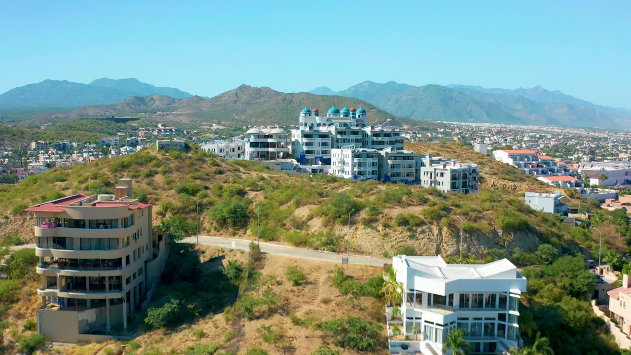 Opening San Jose del Cabo aerial frame with a beige round-front building, white hillside homes, a winding road, dry grass, distant town blocks, mountains, and no visible people.