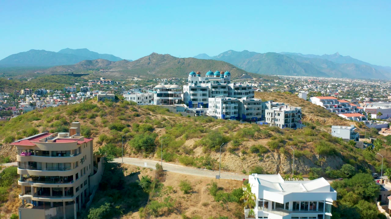 San Jose Del Cabo aerial frame centers more of the hillside homes, including blue-domed roofs, dry slopes, town buildings, distant mountains, and no visible people.