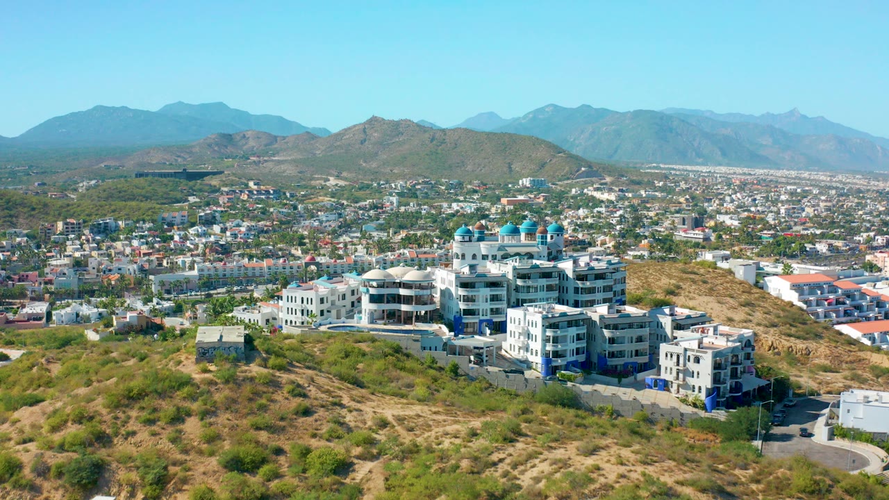 Opening aerial frame in San Jose Del Cabo, Mexico, showing white hillside buildings with blue domes in the lower center, dry scrubland in front, city blocks behind, distant mountains, and no visible people.