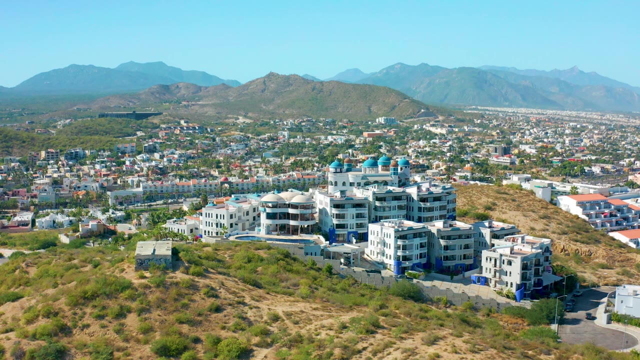 Aerial frame in San Jose Del Cabo, Mexico, looking over dry hillside terrain toward a white building complex with blue domes, dense city blocks beyond, mountain ridges in the distance, and no visible people.