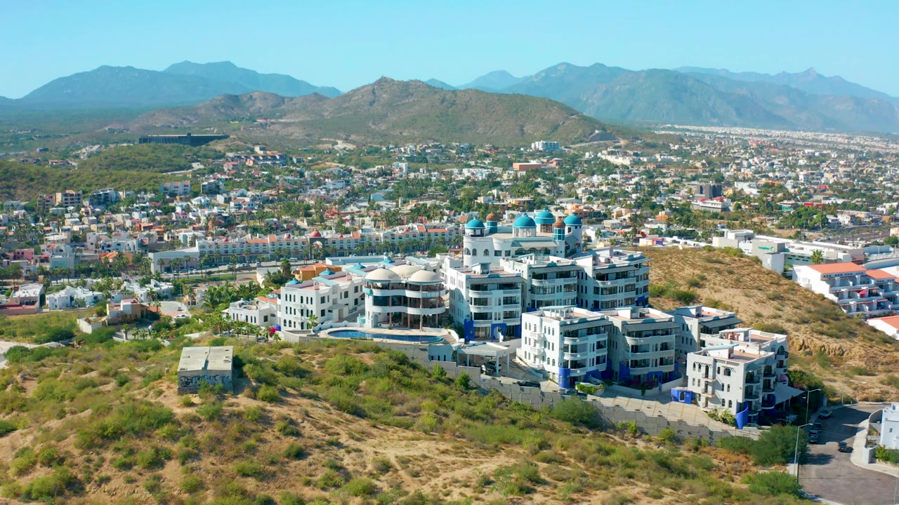 Wide drone view over dry hills, white buildings with blue domes, cityscape, and mountains.