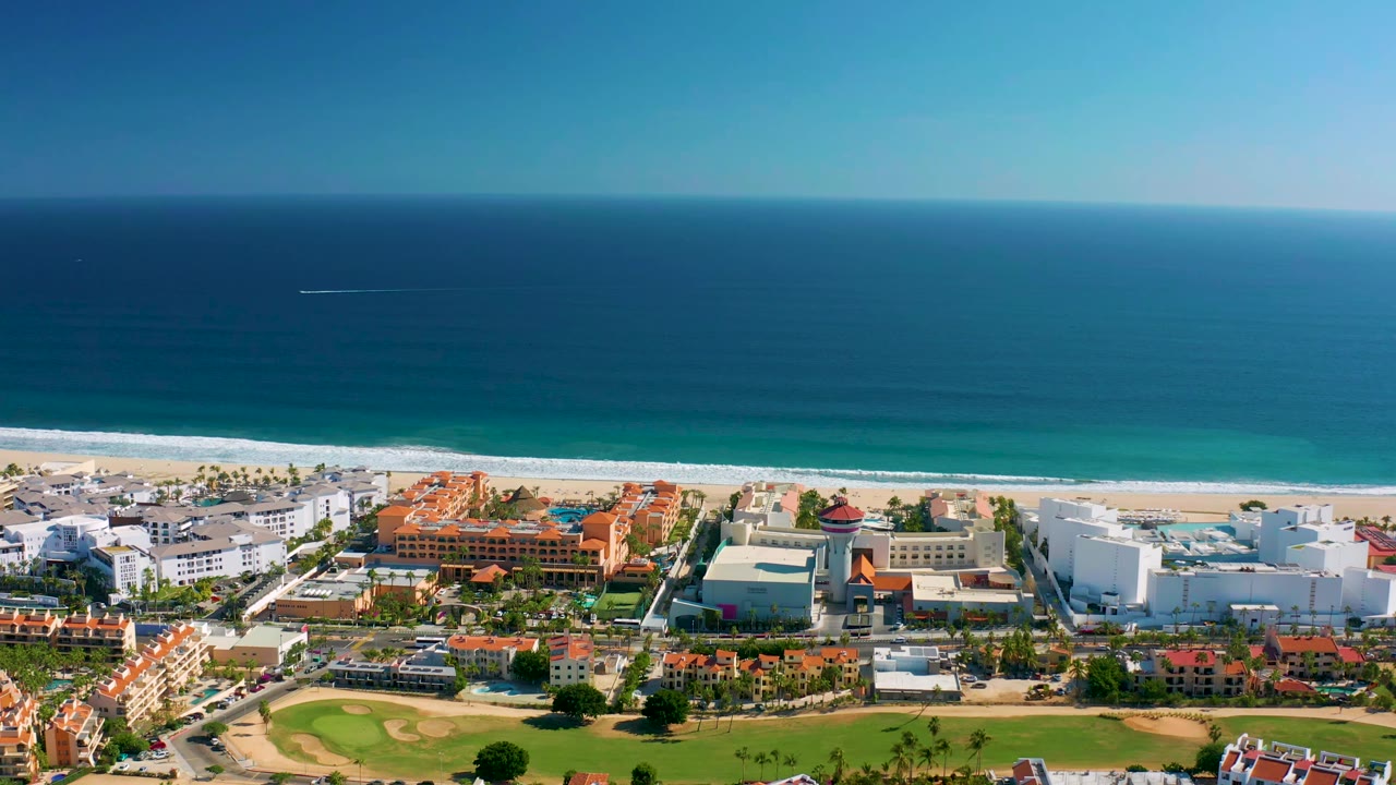 San Jose Del Cabo aerial frame showing the ocean horizon above the beach, white resort structures, red-roofed buildings, and green landscaped areas, with no visible people.