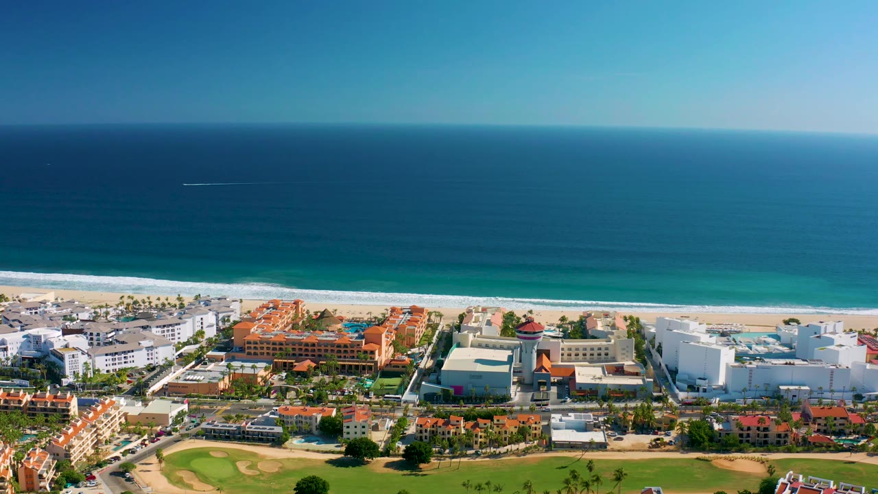 Aerial view in San Jose Del Cabo with the blue ocean filling the background, a pale beach strip, red-roofed resort buildings, white hotel blocks, and no visible people.