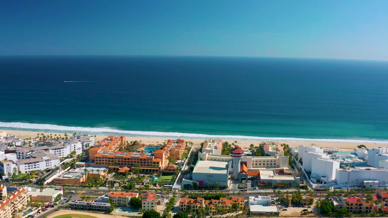 Wide aerial coastline with ocean horizon, beach waves, and beachfront buildings.