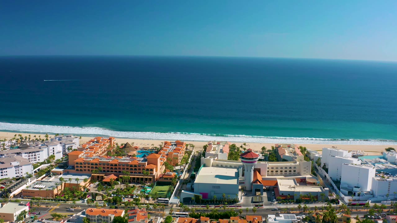 Aerial view in San Jose del Cabo showing white wave foam approaching the sand behind resort-style buildings, a circular tower, and no visible people.
