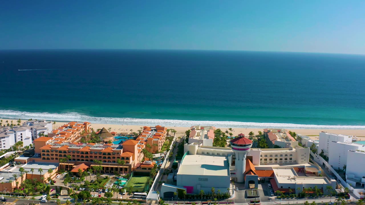 Aerial view in San Jose Del Cabo, Mexico with the ocean horizon high across the frame, white surf meeting sand, orange resort roofs, white buildings, and no visible people.
