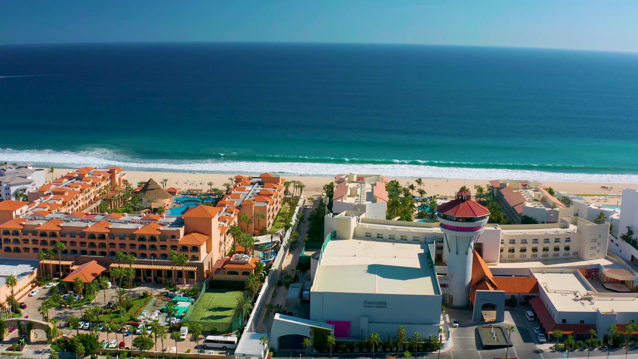 Aerial view in San Jose Del Cabo, Mexico with orange-red resort roofs, blue pools, palm landscaping, a white building, surf line, broad ocean, and no visible people.