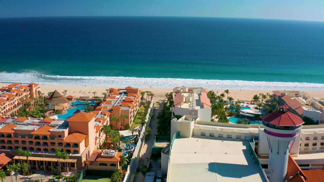 Aerial view over San Jose del Cabo resort rooftops and pools toward a sandy beach and blue ocean, with no visible people.