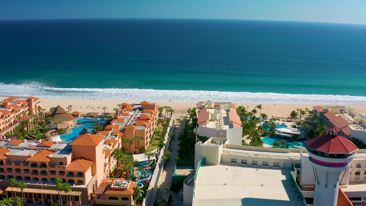 Elevated San Jose del Cabo resort view showing pool courtyards, palm rows, beach frontage, and open blue water with no visible people.