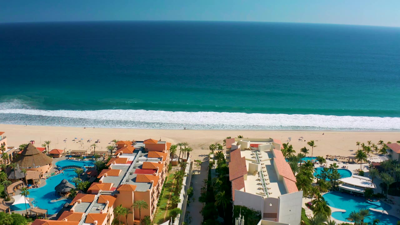 Aerial San Jose del Cabo resort view with red-roof buildings, curved blue pools, palms, sandy beach, and a band of white surf under a clear ocean horizon, with no visible people.