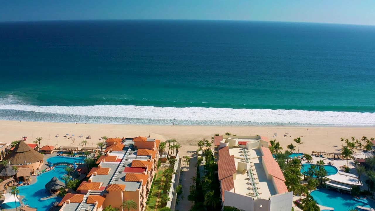 High aerial view over San Jose del Cabo with the ocean filling the upper frame, white waves reaching the beach, and resort pools, palms, and red roofs below, with no visible people.