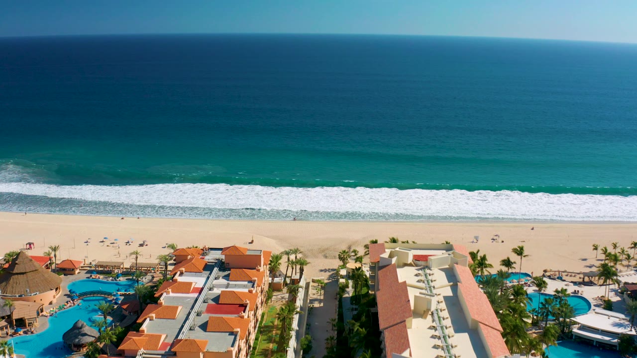 San Jose del Cabo aerial frame showing turquoise nearshore water, a white wave band, pale sand, and resort buildings with pools and palms along the lower edge, with no visible people.