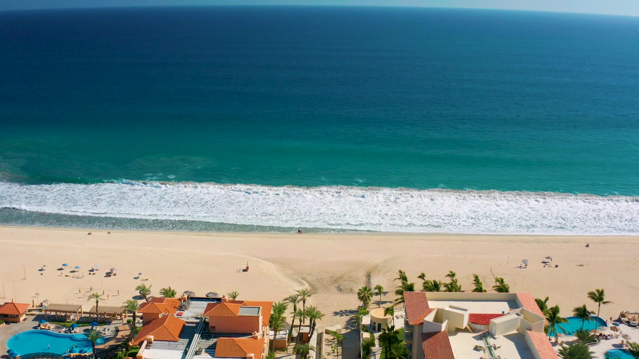 Aerial view in San Jose Del Cabo showing turquoise ocean above a white surf line, pale sand, small beachgoers, umbrellas, palm trees, pools, and resort roofs along the bottom edge.