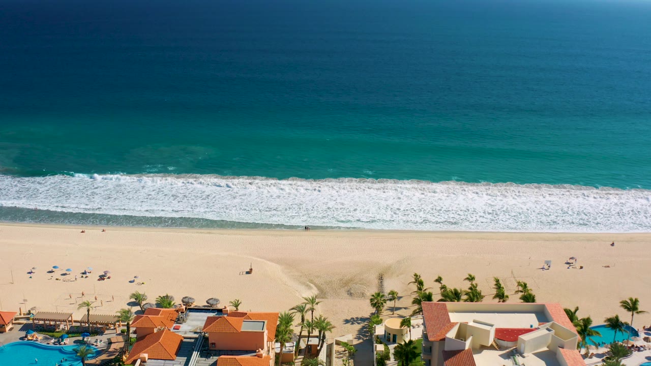 Aerial San Jose Del Cabo beach frame with teal water at the top, foamy surf nearing the sand, sparse beachgoers and umbrellas across the beach, and palm-lined resort grounds below.