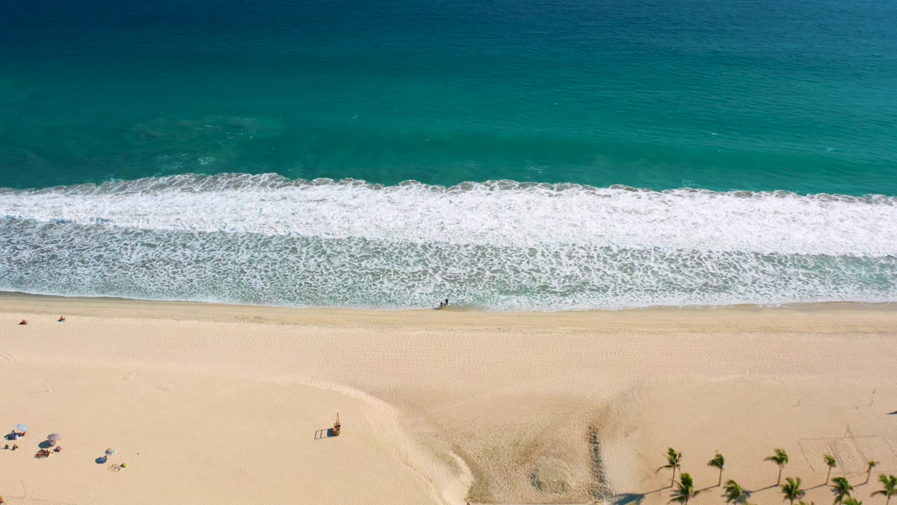 Overhead San Jose del Cabo beach frame showing turquoise ocean, a wide white surf line, pale sand, umbrellas, palm trees near the lower edge, and tiny distant people.