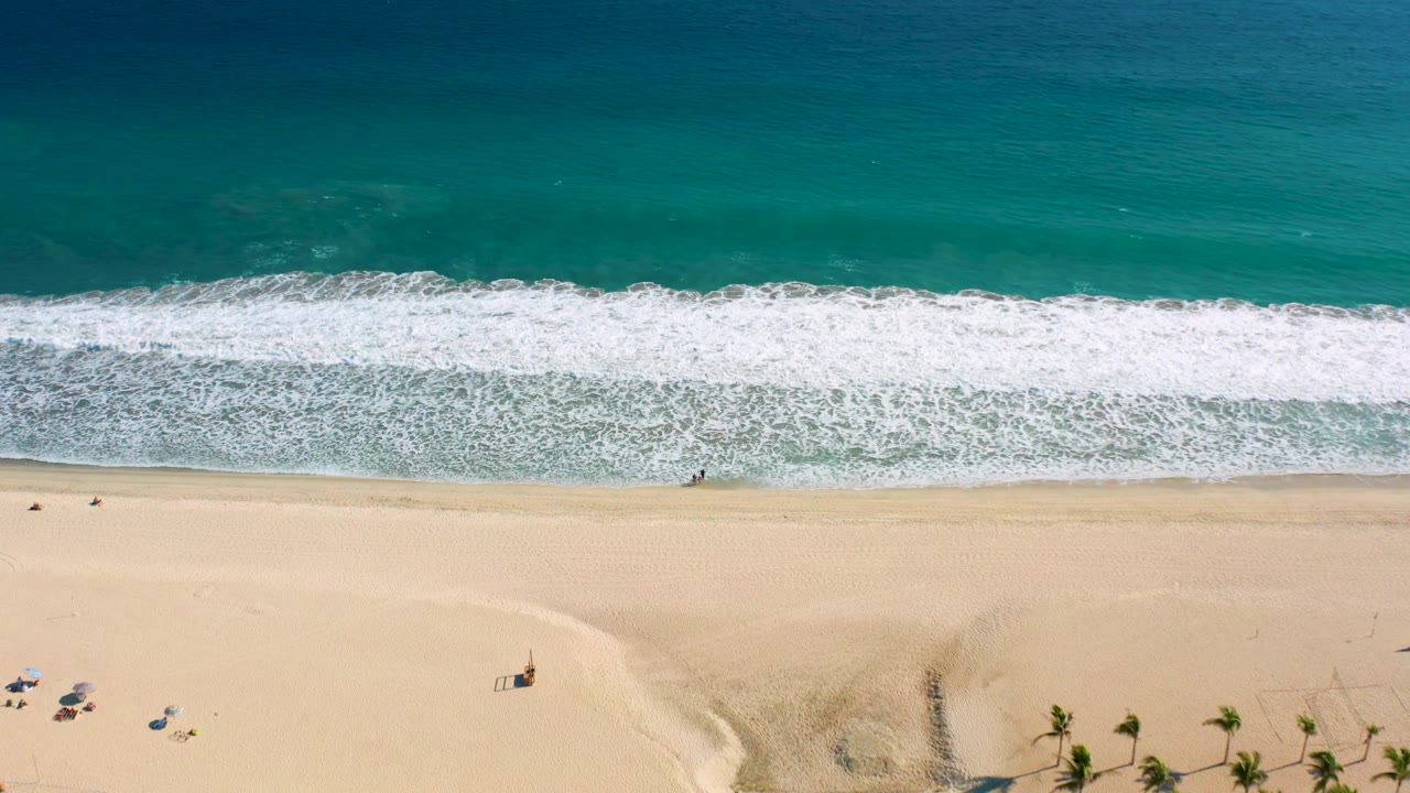 Aerial shoreline with foamy waves, pale sand, umbrellas, palms, and small figures on the beach.