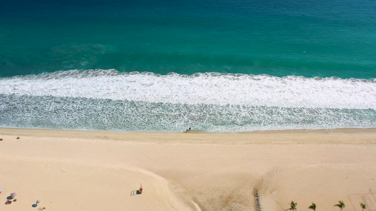 Overhead San Jose del Cabo beach frame with the surf band across the middle, umbrellas and palms low in frame, and tiny people spaced along the sand and waterline.