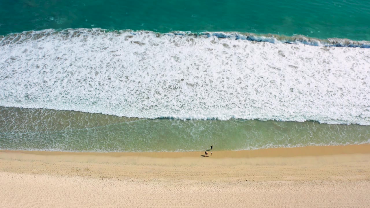 White surf spreads across shallow water above a sandy beach with two small figures below.