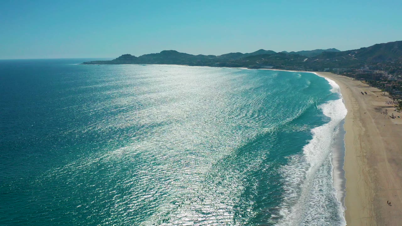 High aerial view over San Jose del Cabo coastline with sunlit blue water on the left, a sandy beach on the right, white surf lines, distant hills, and no visible people.