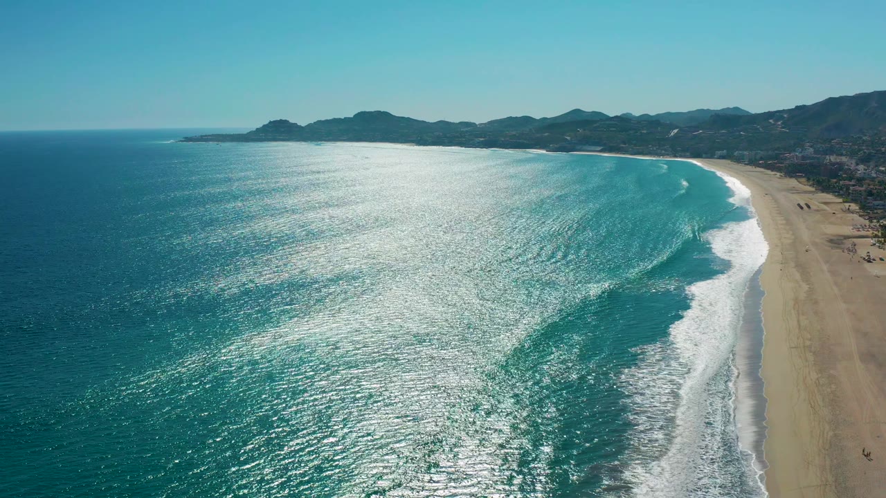 San Jose del Cabo aerial view with turquoise water occupying most of the frame, a narrow strip of beach at right, white foam tracing the shore, distant hills, and no visible people.