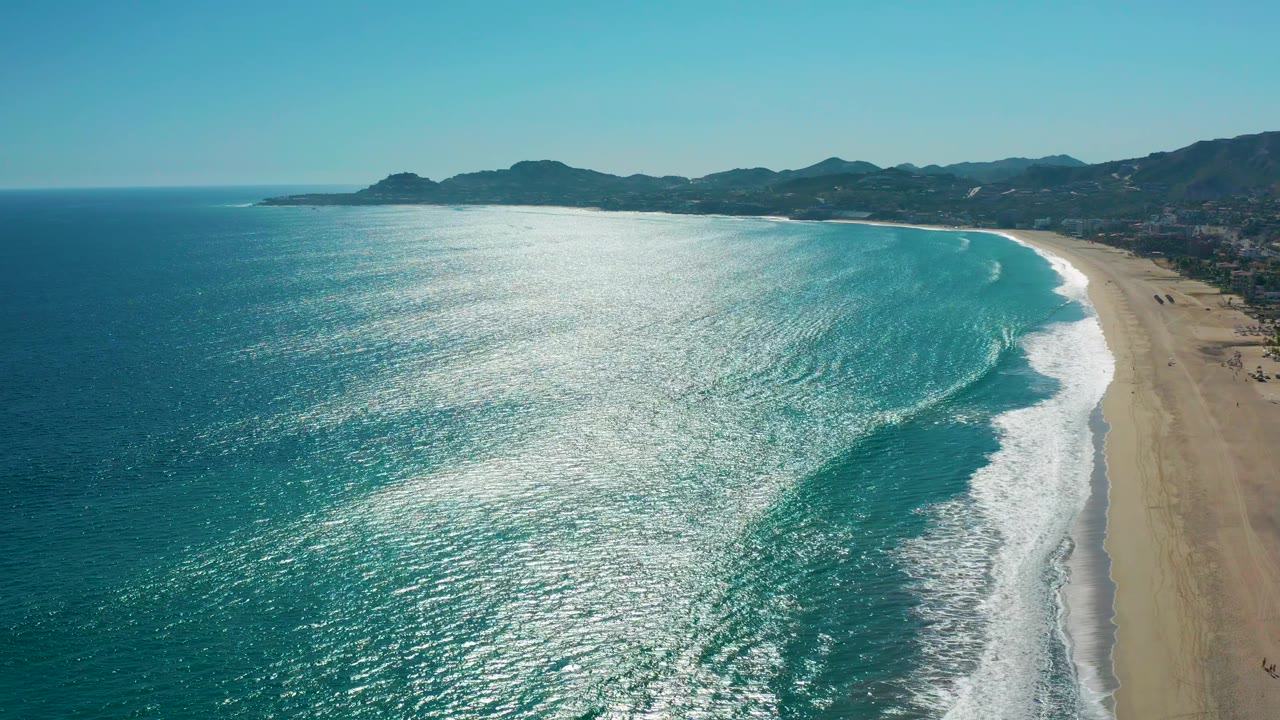 High drone perspective over San Jose del Cabo surf with bright sun reflection across the ocean, white waves curling toward the sandy beach, dark hills inland, and no visible people.