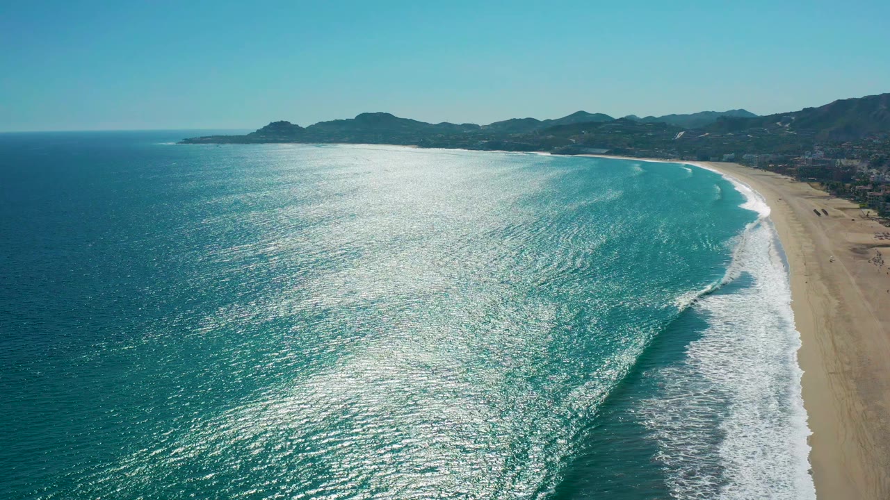 Aerial San Jose del Cabo beach frame with a broad band of sunlit ocean, white wave foam angled toward shore, tan sand on the right, mountains on the horizon, and no visible people.
