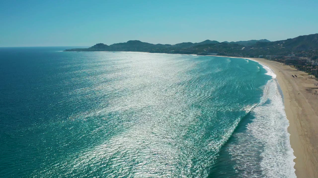 Elevated view of the San Jose del Cabo shore with the reflective ocean filling most of the frame, a curved white surf break near the beach, background mountains, and no visible people.