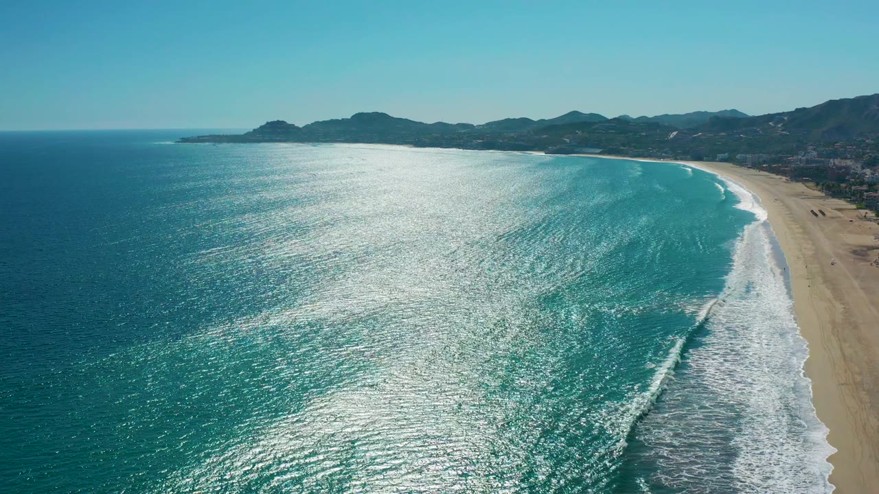 San Jose del Cabo aerial coastline with deep blue water at left, bright sun glare across the midframe, turquoise shallows near the right-side beach, white foam, and no visible people.