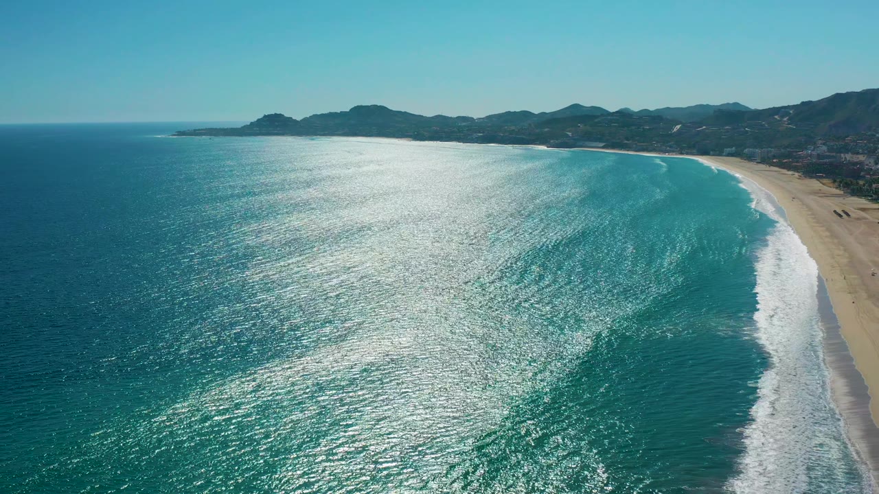 Wide aerial frame of San Jose del Cabo coastline with a long white wave band meeting the sandy beach, teal water in the foreground, mountains in back, and no visible people.